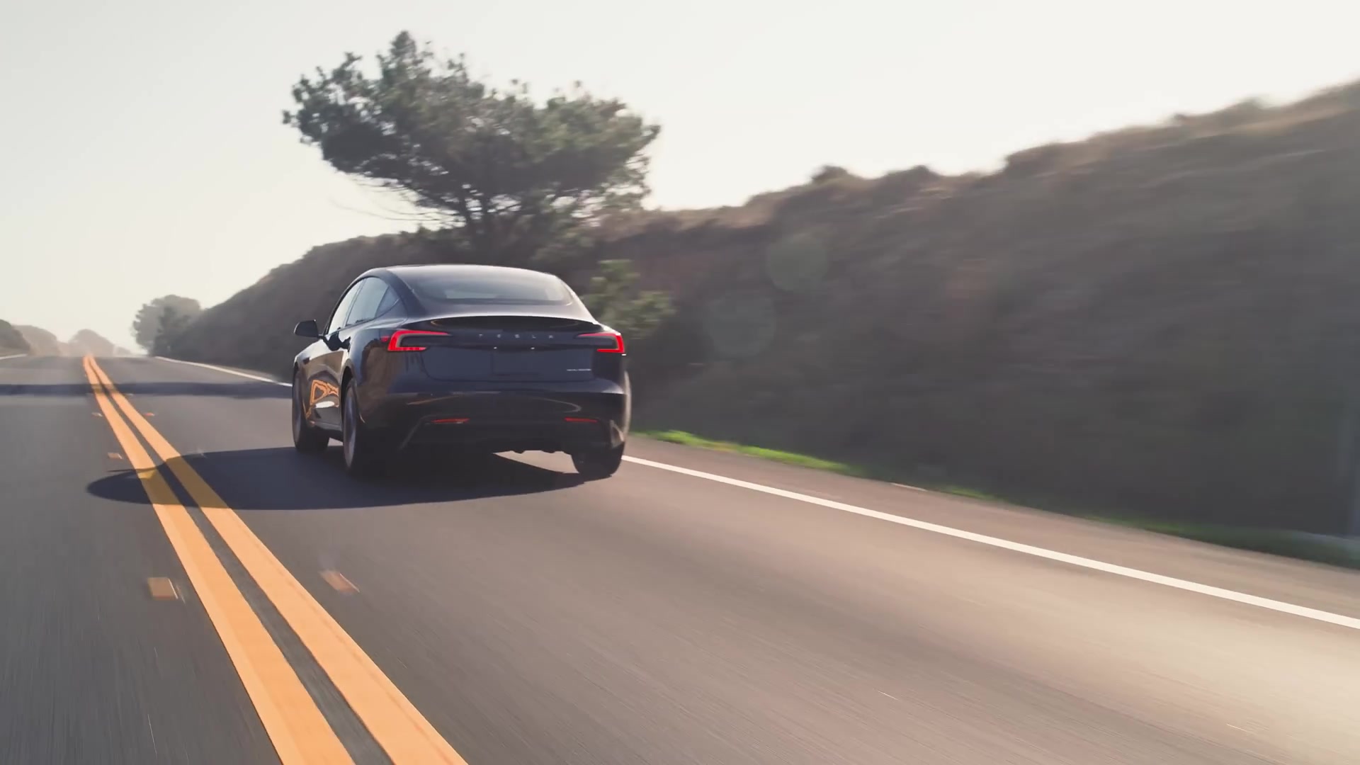 Donkere Tesla Model 3 rijdt over een tweebaansweg langs een heuvel